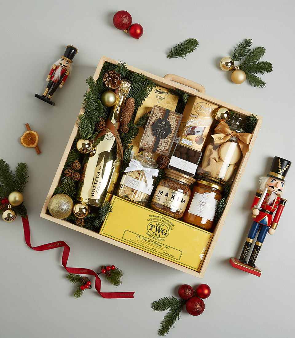 Festive wooden crate with gold prosecco, gourmet tea, preserves, and chocolates decorated with pine and golden baubles for