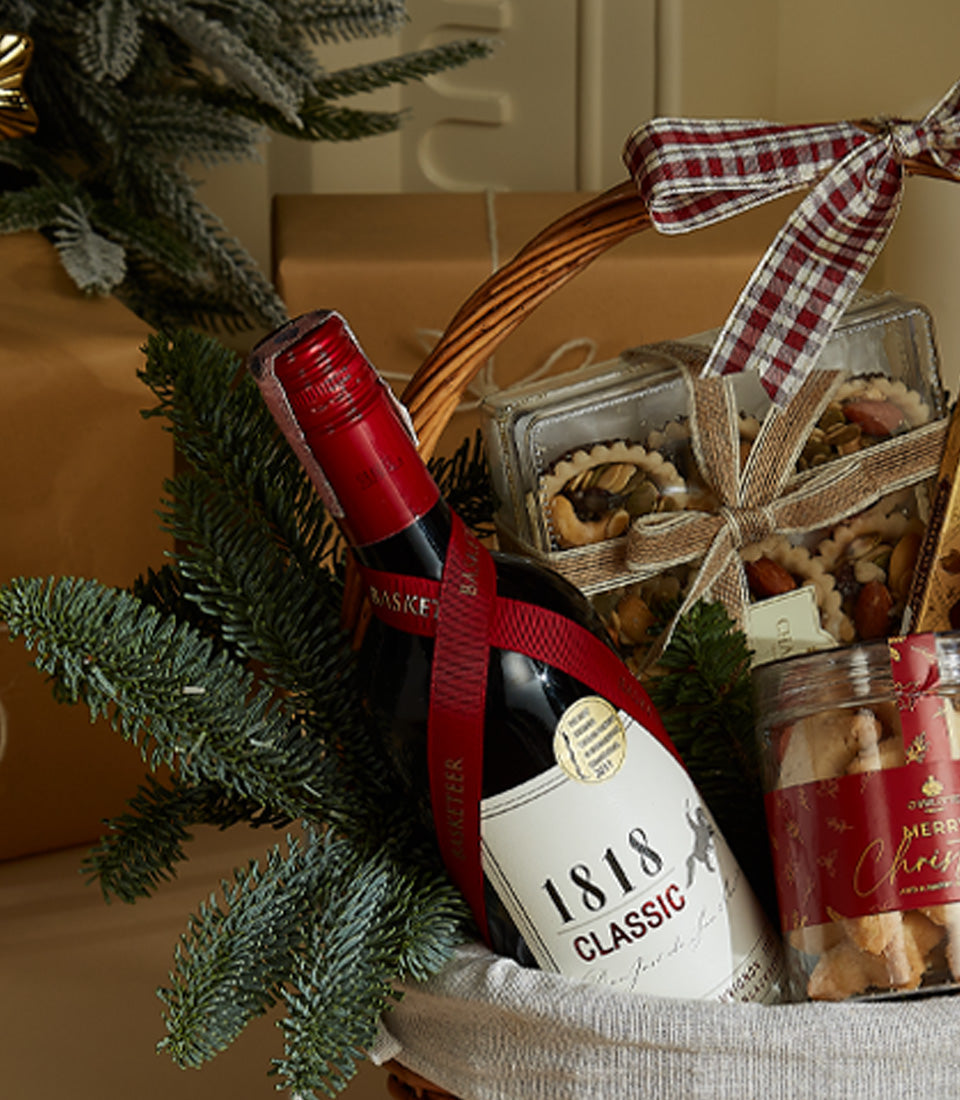 Premium red wine gift basket with gourmet tarts and holiday cookies, tied with festive ribbons among evergreen branches