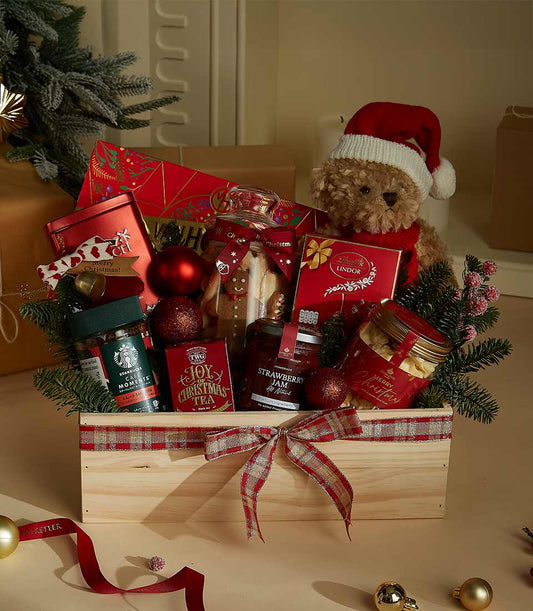 Festive wooden gift hamper with teddy bear, gourmet chocolates, cookies, jam, coffee, and tea among pine branches and red