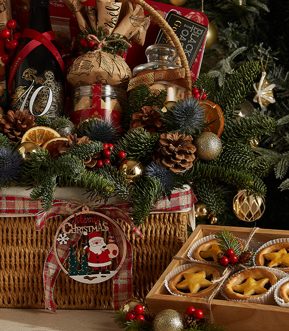 Wicker Christmas hamper with red plaid ribbon, red wine, cookies, and festive pine decorations alongside star-shaped tarts
