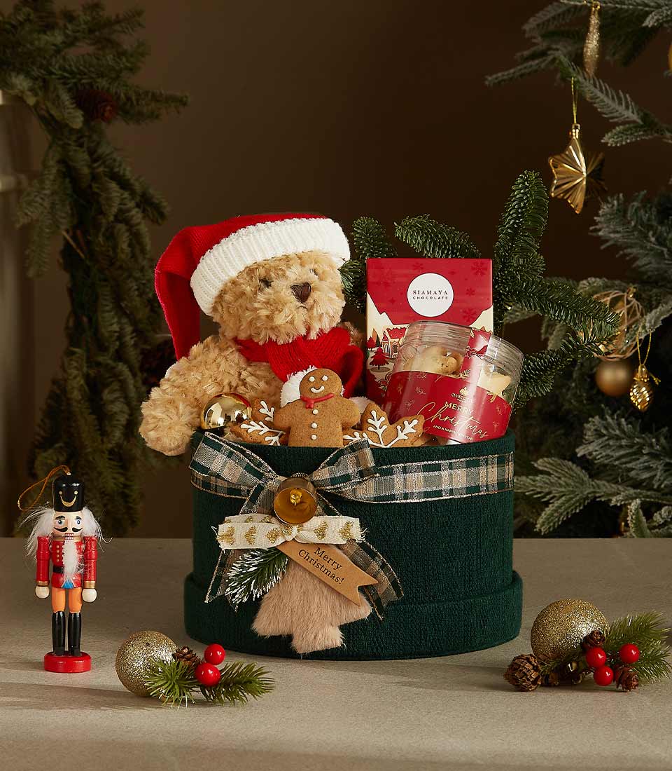 Dark green cylindrical Christmas gift box with plaid ribbon, stuffed teddy bear in Santa hat, artisan chocolates, and