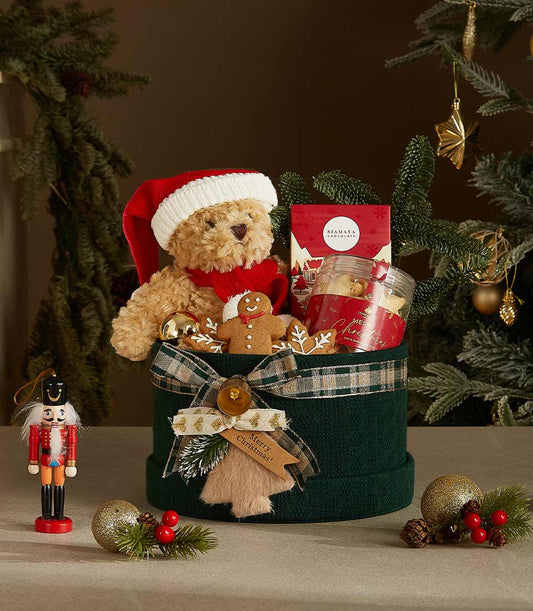 Dark green cylindrical Christmas gift box with plaid ribbon, stuffed teddy bear in Santa hat, artisan chocolates, and