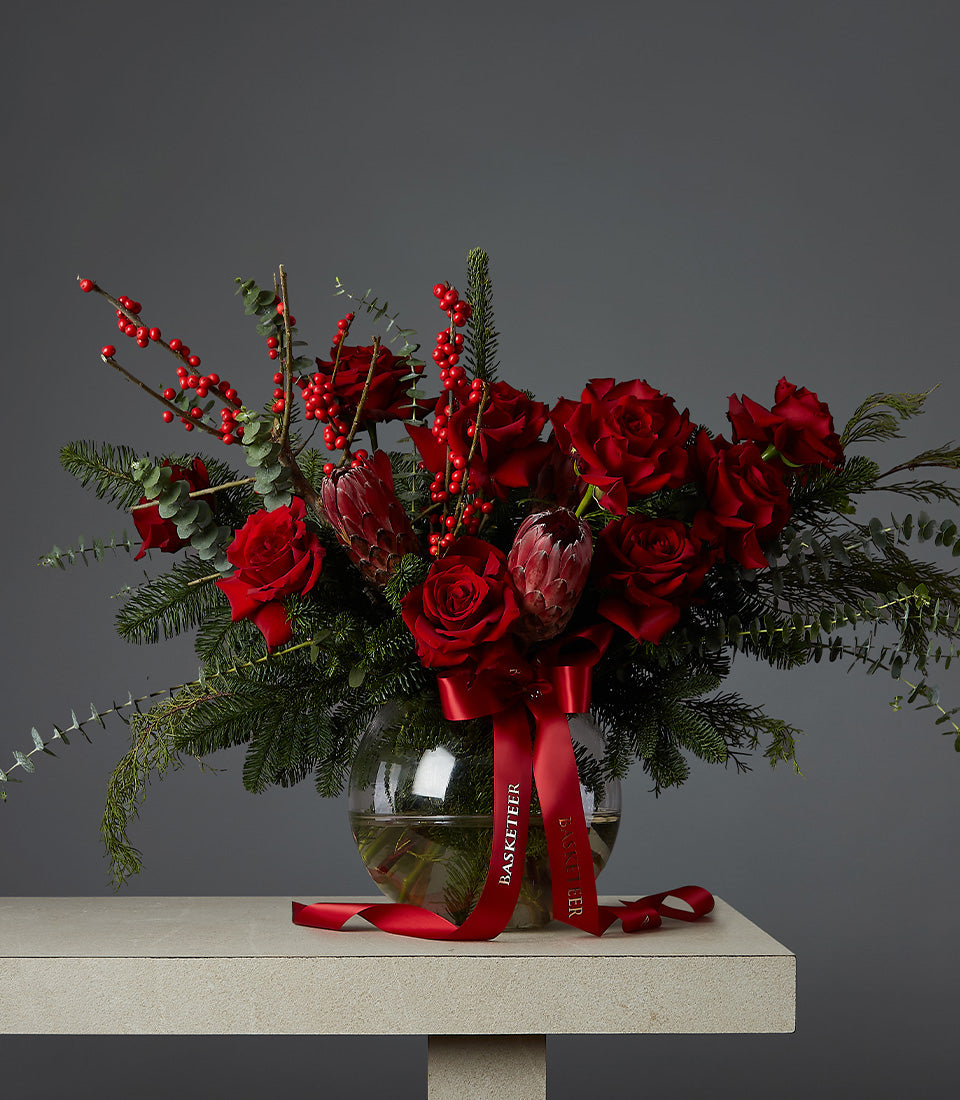 Crimson festive rose arrangement with red roses, protea, and pine in glass vase with red ribbon, perfect for Christmas