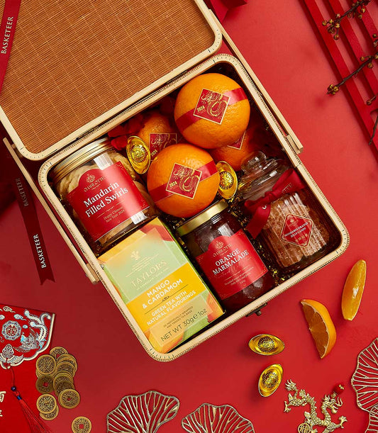 Basketeer Lunar New Year gift in woven basket: oranges with festive stickers, mandarin swirl cookies, marmalade, and aromatic tea. Perfect premium hamper delivery in Thailand.