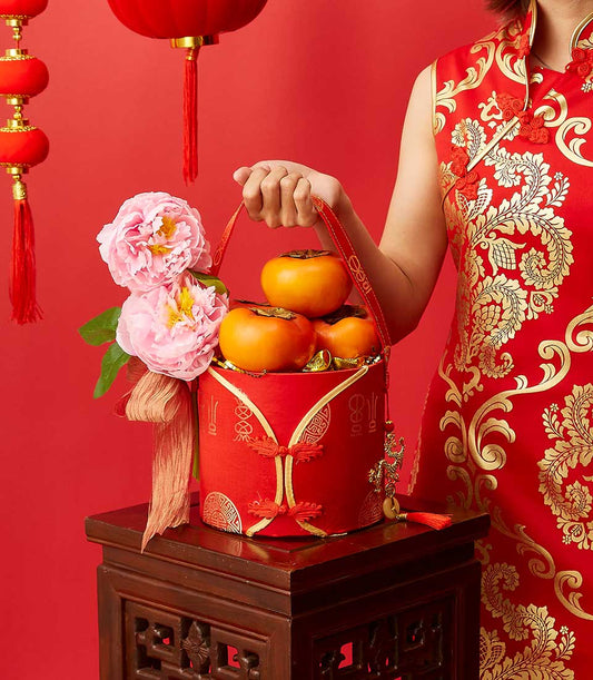 Lunar New Year gift box with persimmons and gold ingots, pink peonies, traditional red cheongsam styling on carved wooden
