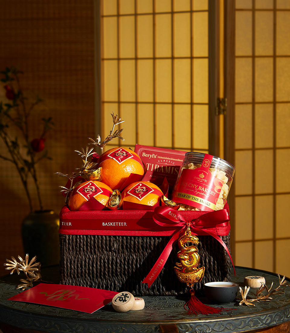 Basketeer premium hamper delivery Thailand: Oranges, sweets, & chocolates artfully arranged in a dark wicker gift basket with red ribbon. A corporate gift of prosperity.