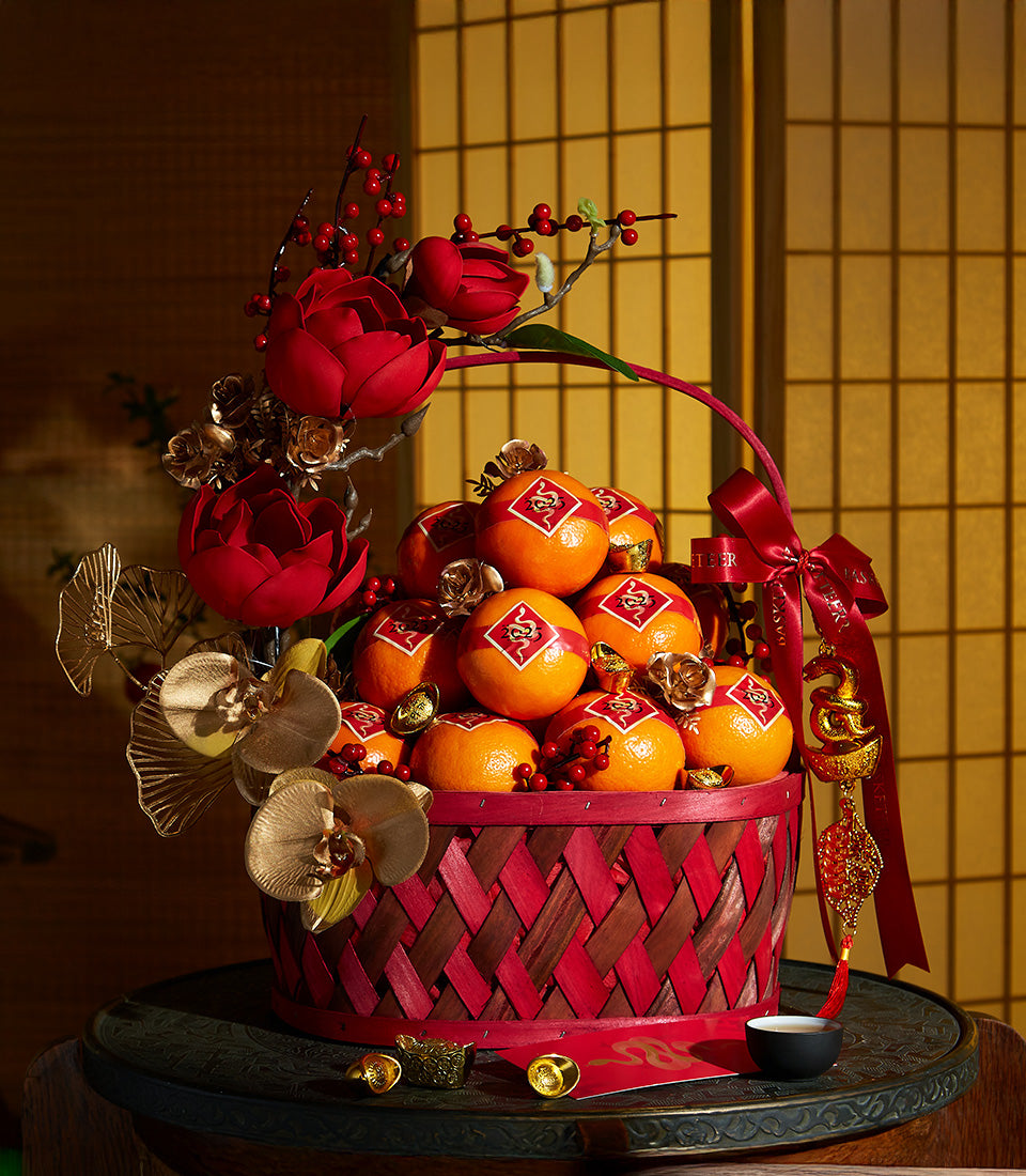 Woven red and gold Lunar New Year gift basket overflowing with fresh oranges, silk flowers, and prosperity charms