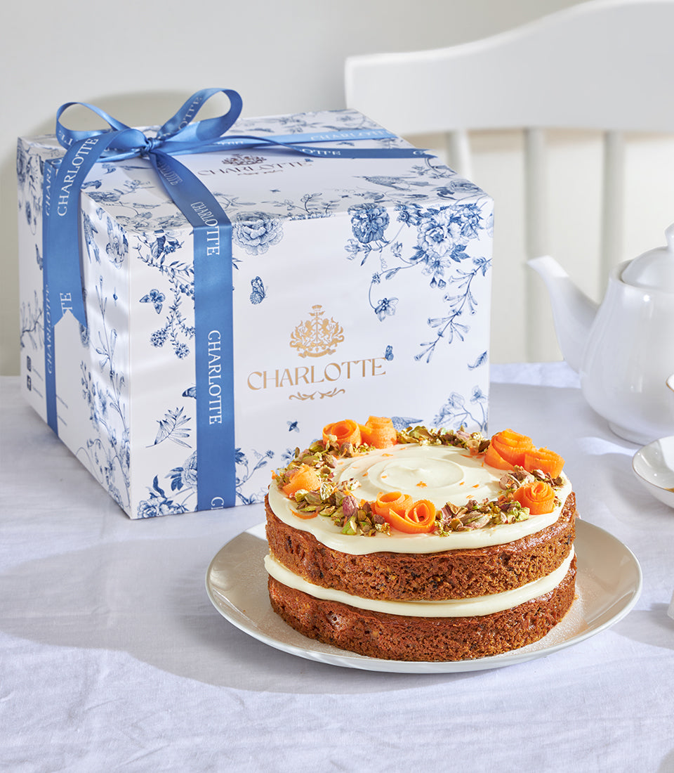 Frosted carrot cake with cream cheese icing and pistachio garnish, presented in Charlotte Bakery gift box with blue floral