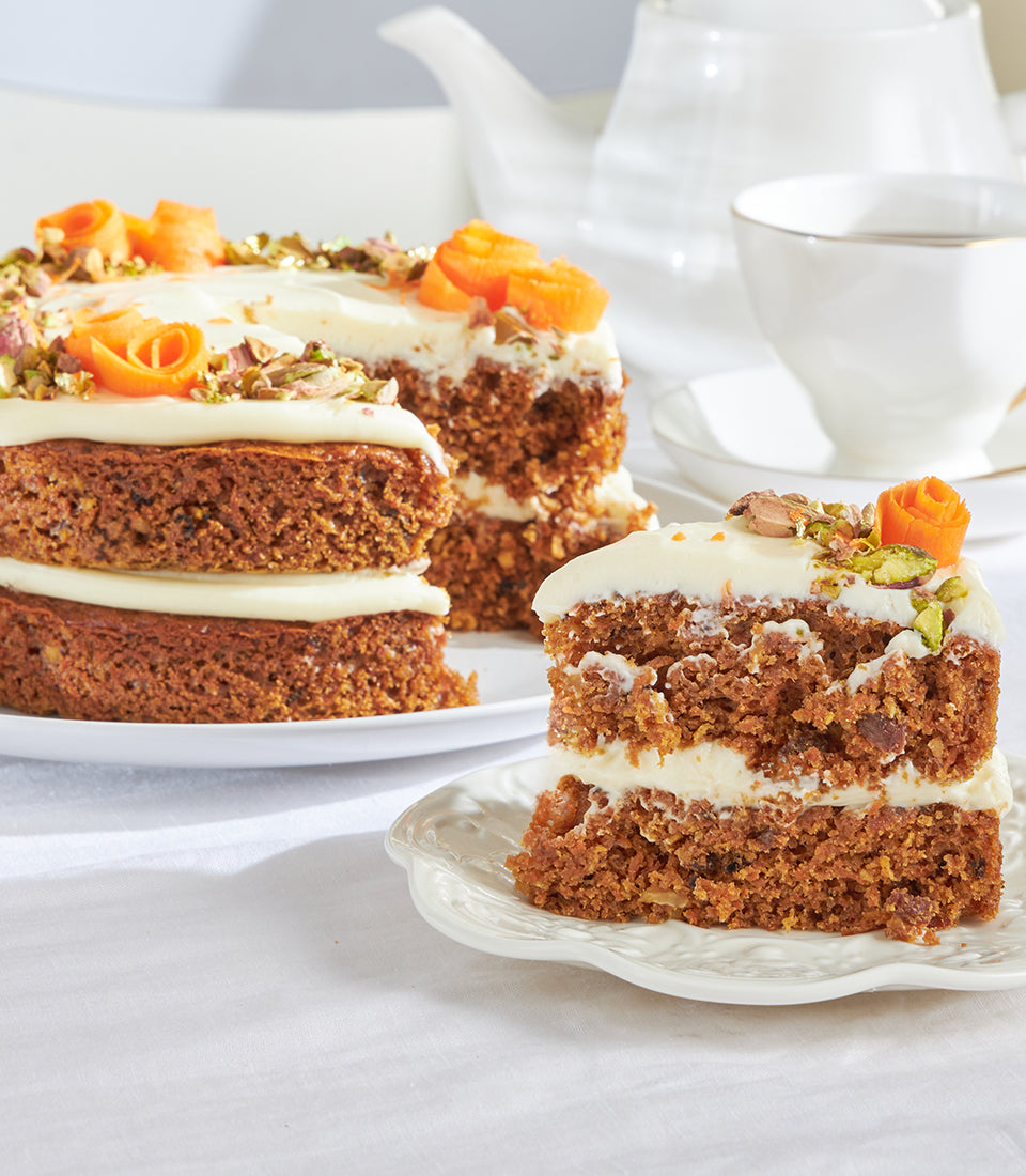 Two-tiered carrot cake with cream cheese frosting, candied carrot curls, and pistachio garnish, served with tea