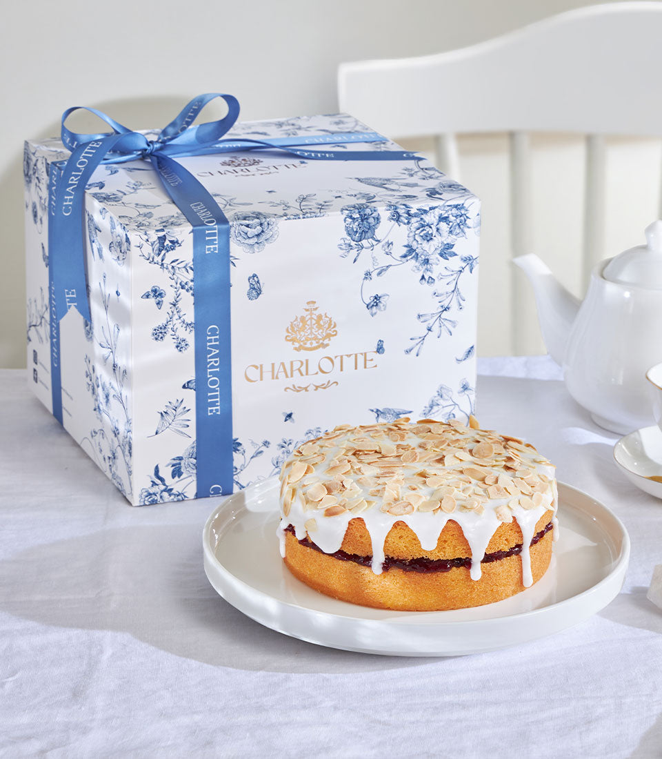 Cherry Bakewell almond cake with white icing drip and sliced almonds, presented in blue floral gift box with satin ribbon