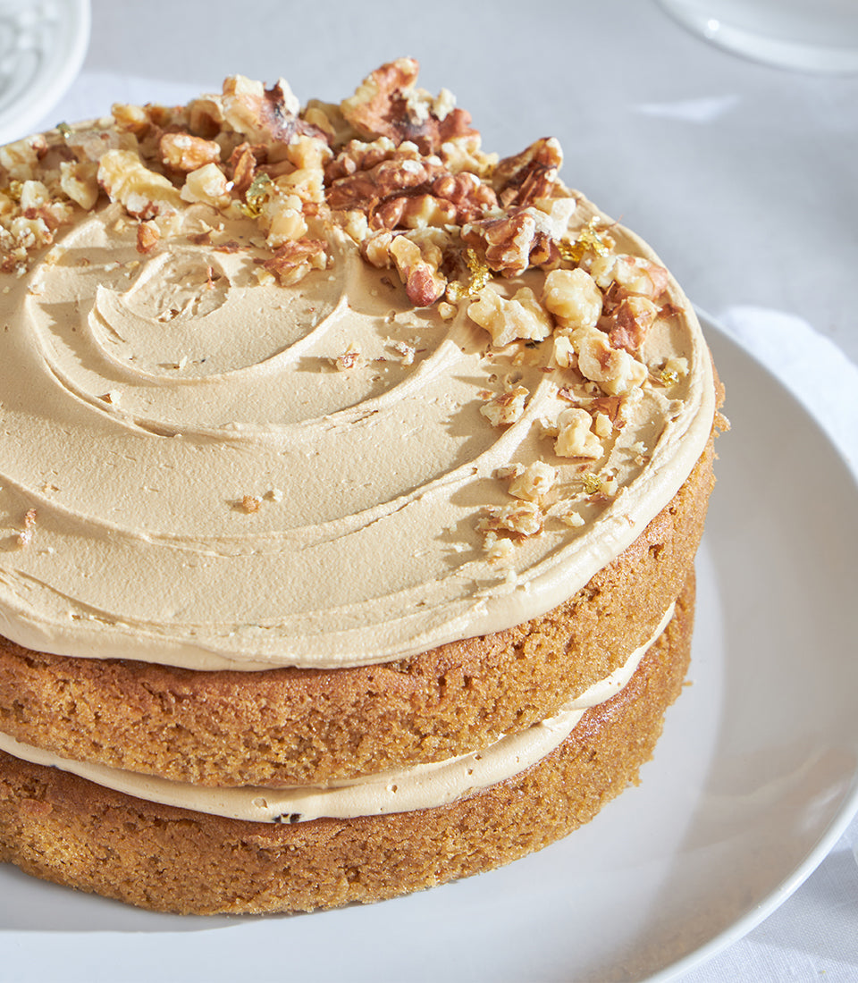 Two-layer coffee walnut cake with smooth tan buttercream, walnut crunch, and gold leaf on white plate
