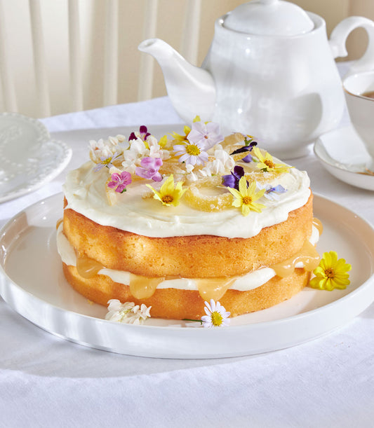 Two-tier lemon drizzle cake with white frosting, caramel glaze, edible flowers and candied lemon on ceramic plate