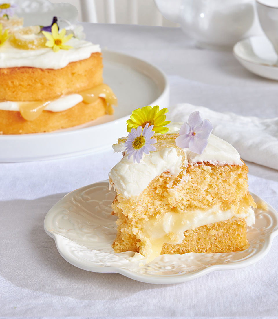 Lemon drizzle cake slice with cream frosting, candied lemon, and edible flowers on white plate