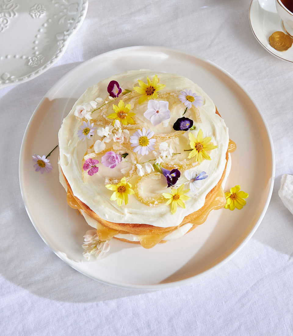 Zesty lemon drizzle cake with white frosting, golden filling, and candied lemon peel flowers on white plate