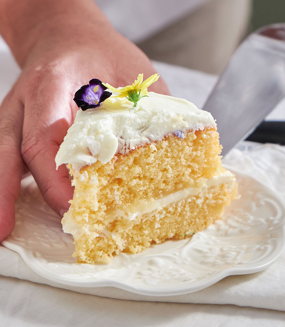 Slice of zesty lemon drizzle cake with white frosting and edible flower garnish on decorative plate