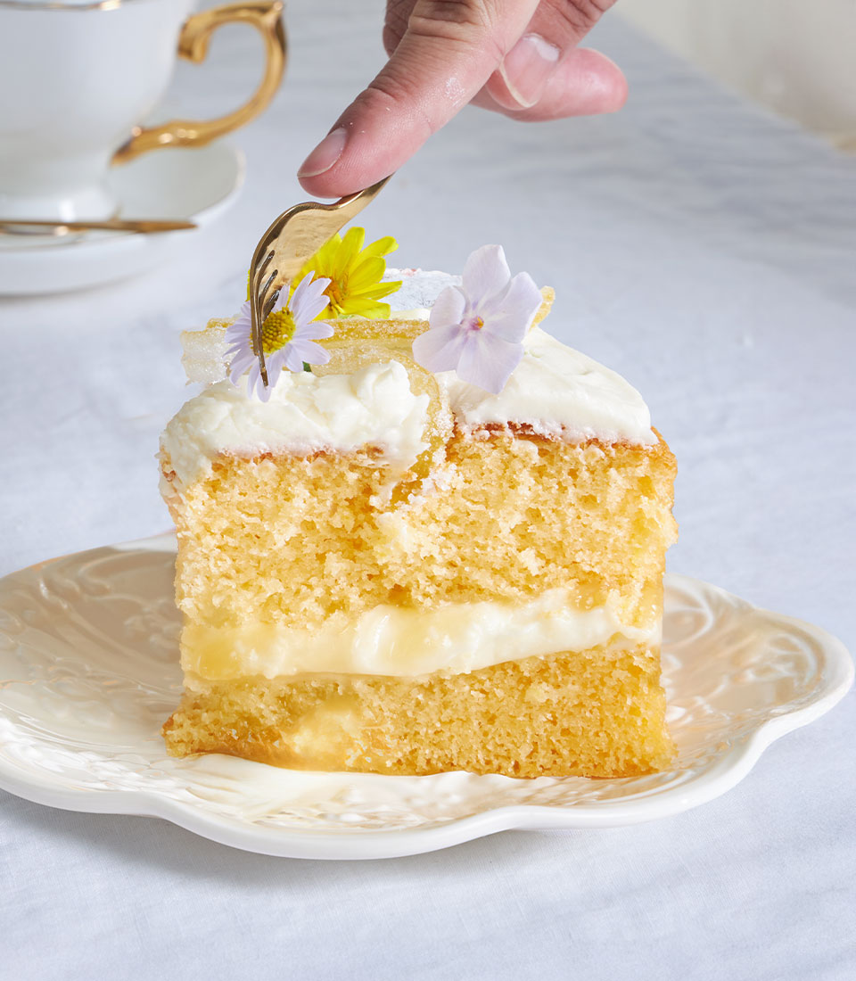 Lemon drizzle layer cake with creamy filling, edible flowers, and gold fork on scalloped plate