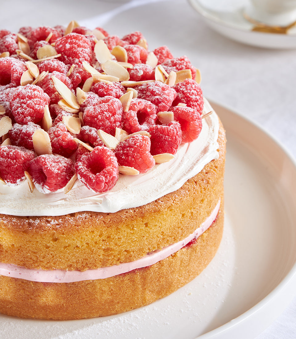 Raspberry and almond celebration cake with fresh berries, whipped cream, and slivered almonds on white ceramic plate