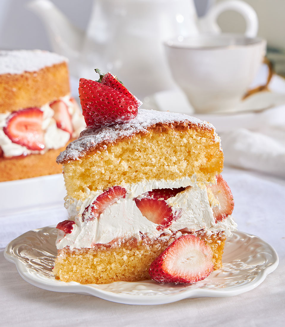 Thai-style strawberry Victoria sponge cake with whipped cream, fresh berries, and powdered sugar served on scalloped plate