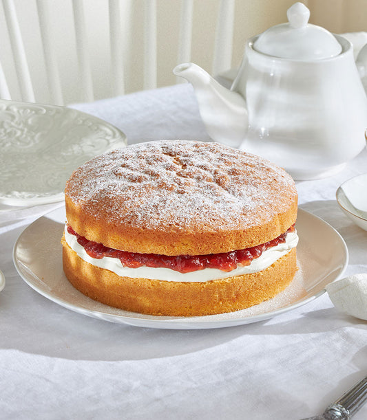Classic Victoria Sponge cake with jam and cream, dusted with icing sugar, presented on white ceramic with teapot