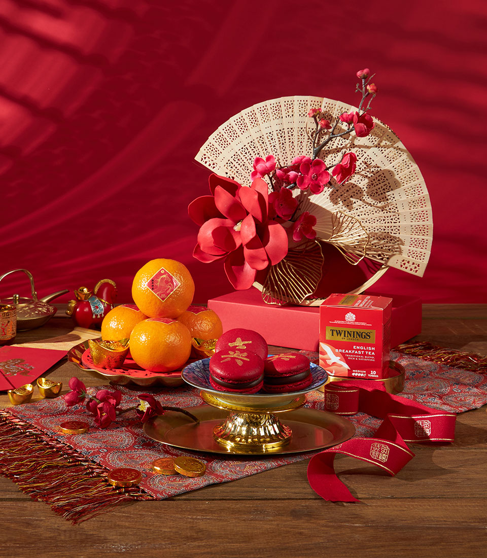 Mandarin, tea and macaron Chinese New Year gift set with red florals, gold accents and ornate cream fan on wooden surface