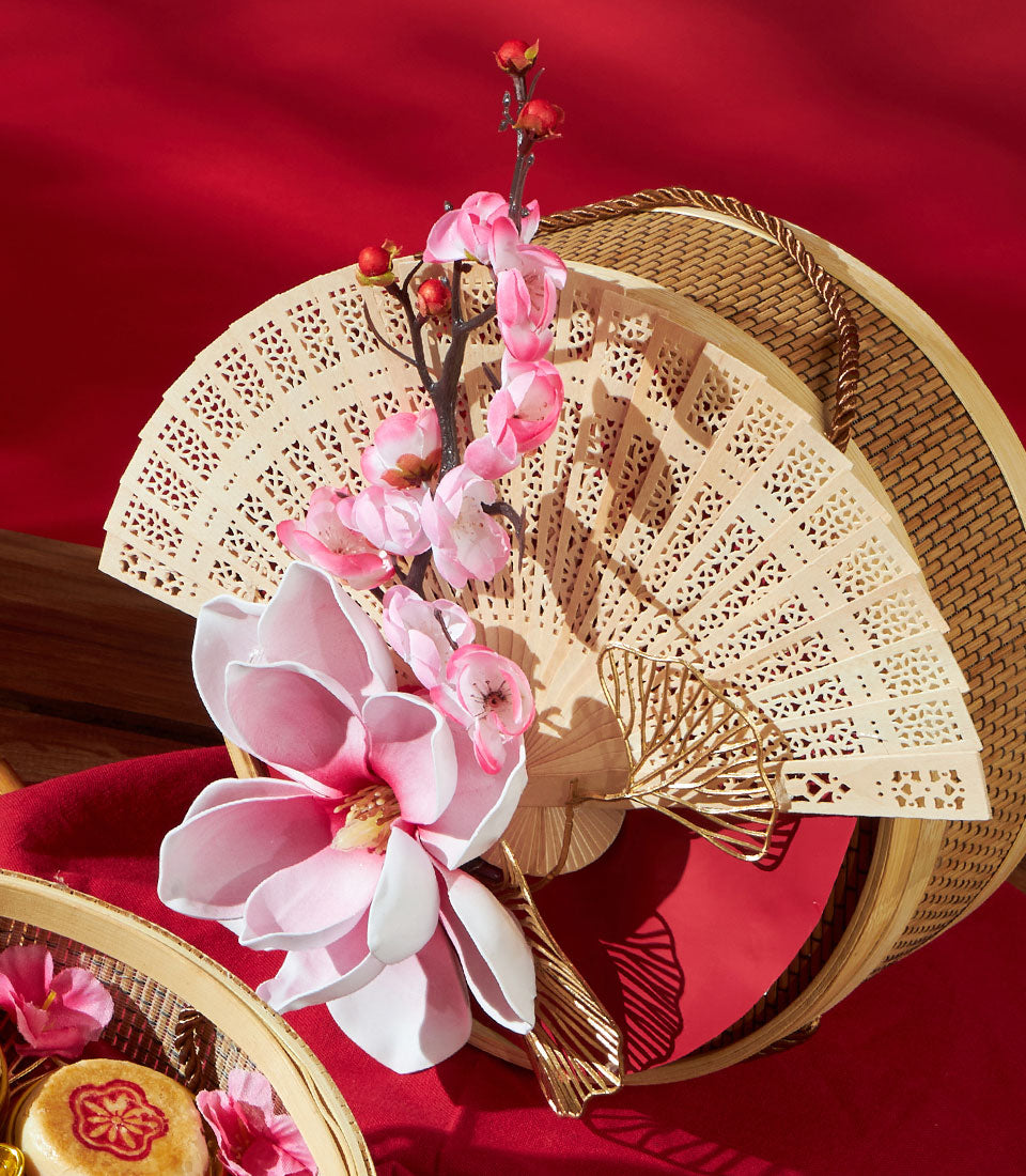 Assorted Chinese pastry gift box with carved wooden fan, silk flowers, and mooncake on festive red backdrop