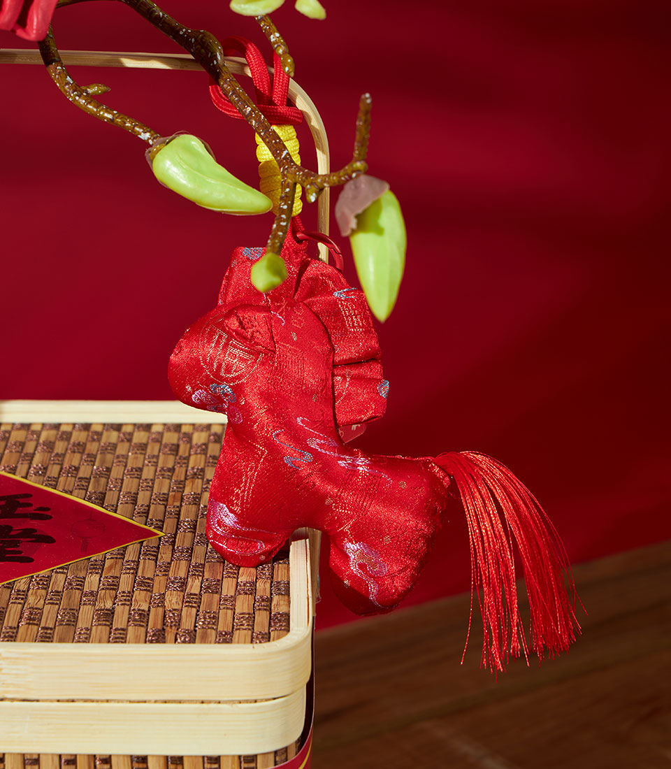 Red fabric horse ornament with tassel, hanging above mandarin oranges and traditional pastry gift box for Chinese New Year