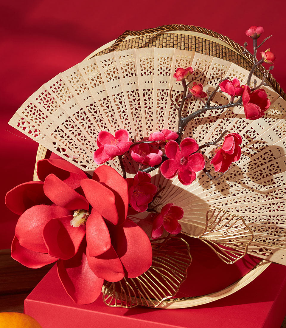 Mandarin and pastry gift set with decorative wooden fan, red flowers, and gold accents on red background for Chinese New Year
