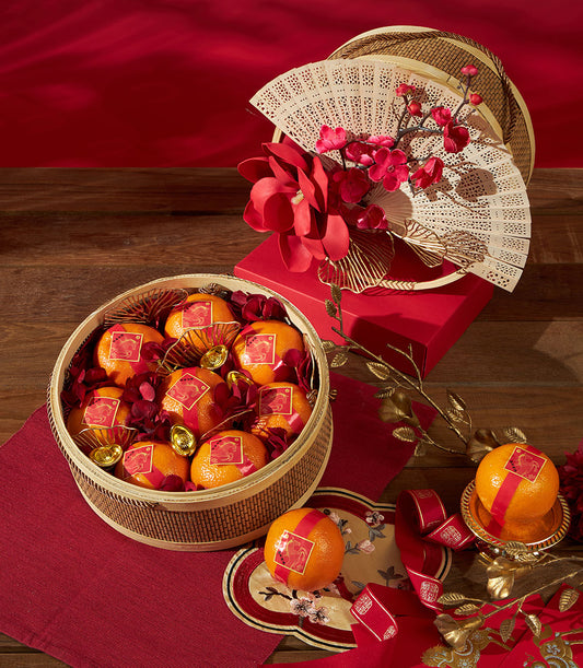 Woven bamboo basket of mandarin oranges with red ribbons and gold zodiac labels, styled with gold ingots and cherry blossoms