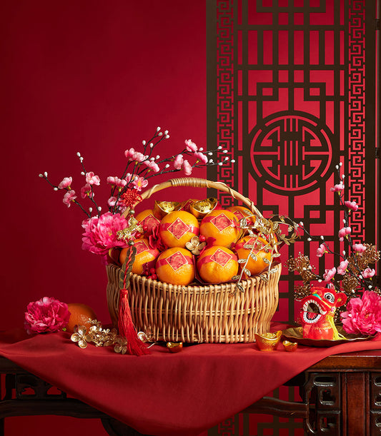 Woven wicker basket filled with red-stickered mandarins, pink peonies, gold ingots, and festive red berries for prosperity