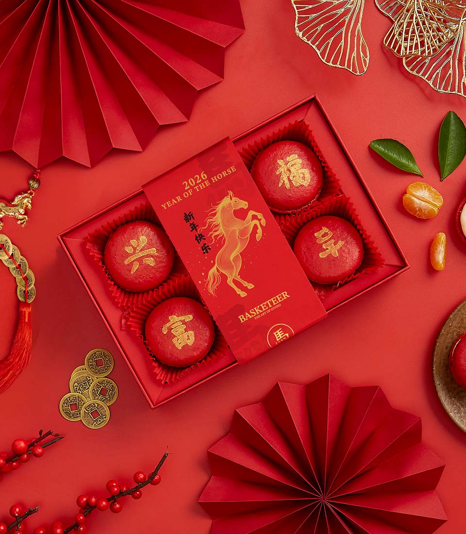 Red macaron gift box with gold Chinese characters for Lunar New Year, surrounded by festive decorations and mandarin oranges