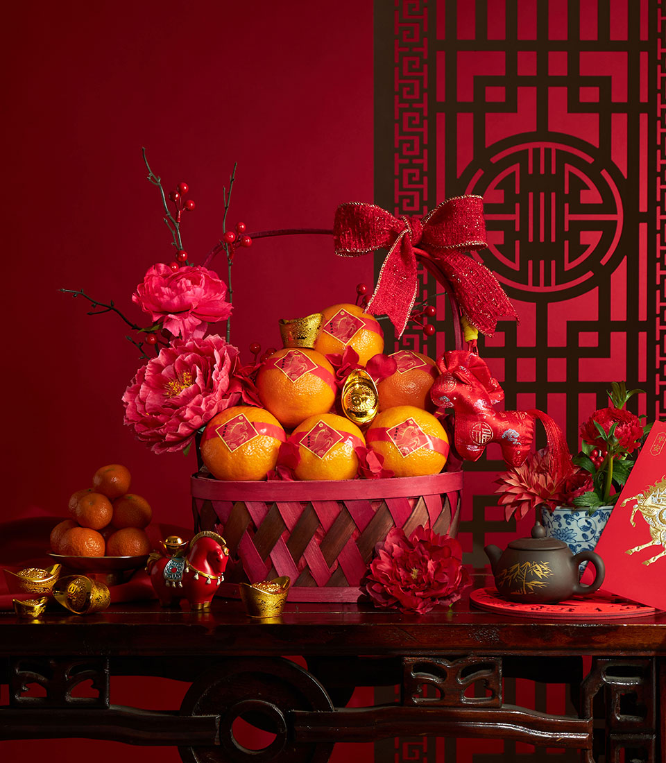 Woven red and brown basket filled with fresh mandarins, decorated with artificial red peonies and festive gold accents for