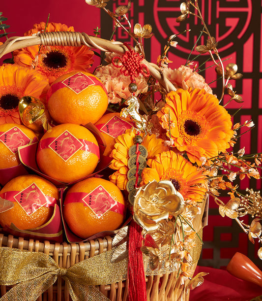Golden wicker basket with mandarin oranges, orange gerberas, and gold-accented florals tied with sheer gold ribbon for