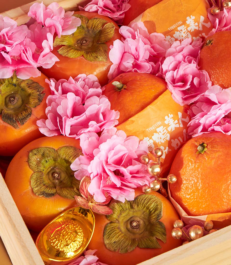 Premium Chinese New Year fruit gift box with persimmons, tangerines, pink blossoms, and gold ornaments in wooden basket