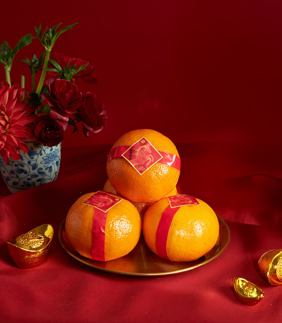 Festive Mandarin orange gift set with golden tray, red ribbons, ceramic vase of dahlias, and traditional wealth symbols for