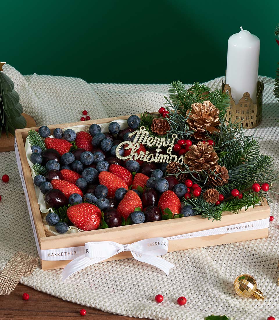 Wooden gift box with fresh strawberries, blueberries, and cherries, festive pine branches, and gold "Merry Christmas"