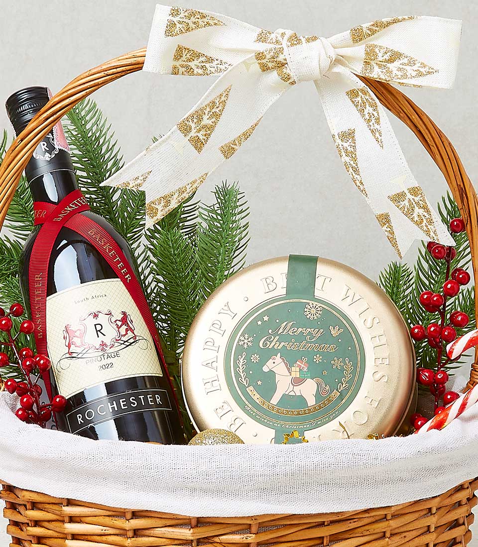 Woven wicker holiday basket with South African Pinotage wine, festive Christmas tin, and seasonal greenery