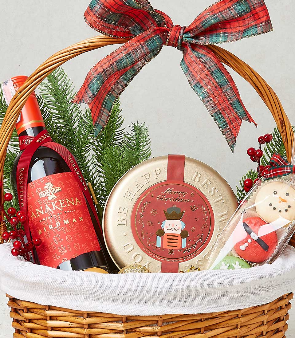 Woven wicker holiday gift basket with red wine, gold Christmas tin, festive cookies, and plaid ribbon