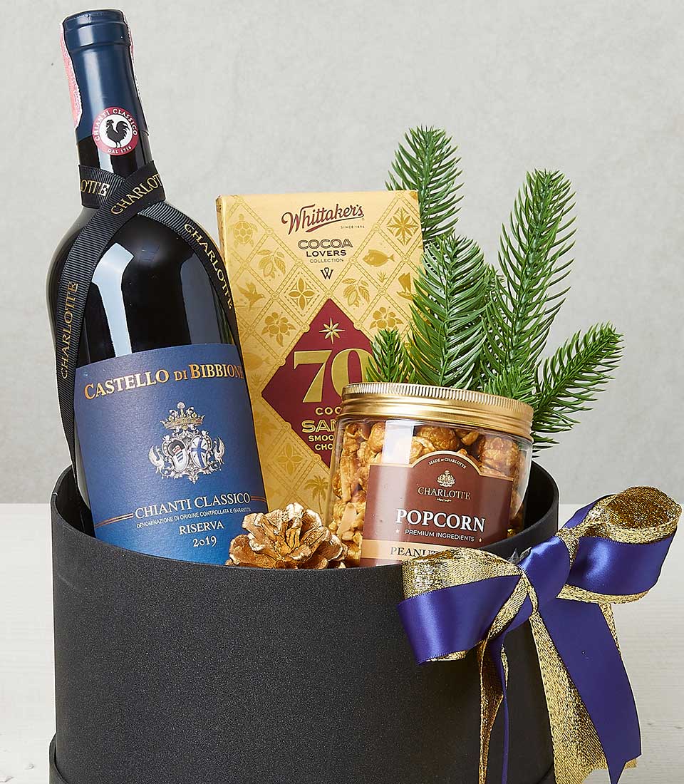 Festive red wine, chocolate, and popcorn gift set in black cylindrical basket with gold pinecone and holiday ribbons