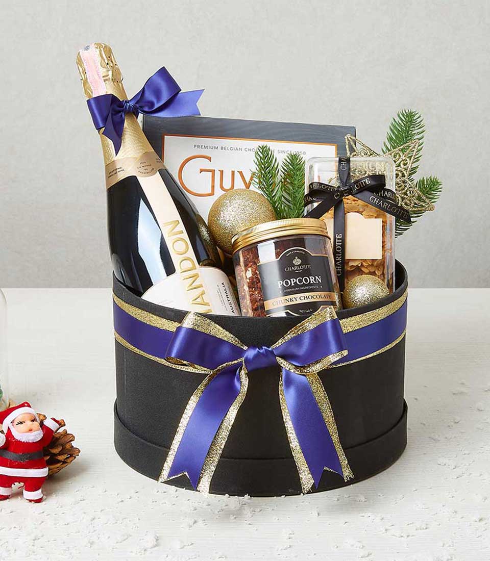 Festive black gift box with sparkling wine, chocolate popcorn, and cookies tied with royal blue ribbon and gold bow