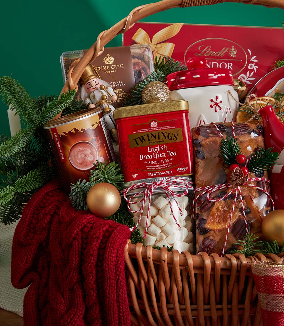 Woven wicker Christmas gift basket with tea, hot chocolate, fruitcake, marshmallows, and luxury chocolates on dark green