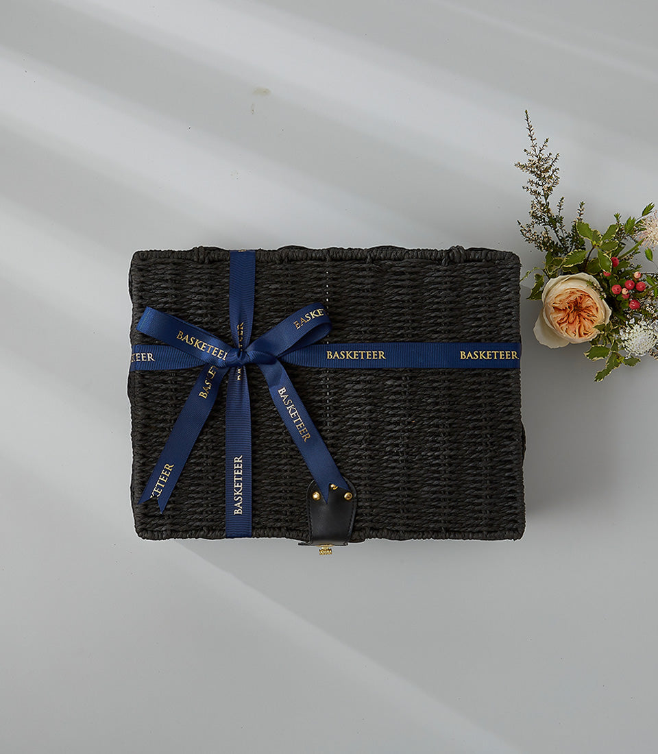 Black wicker gift hamper with leather handles and gold studs, paired with fresh orange rose and berry arrangement