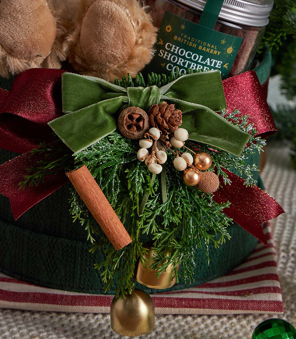 Dark green knitted Christmas gift box with plush velvet bow, pinecones, white berries, gold baubles, and cinnamon stick