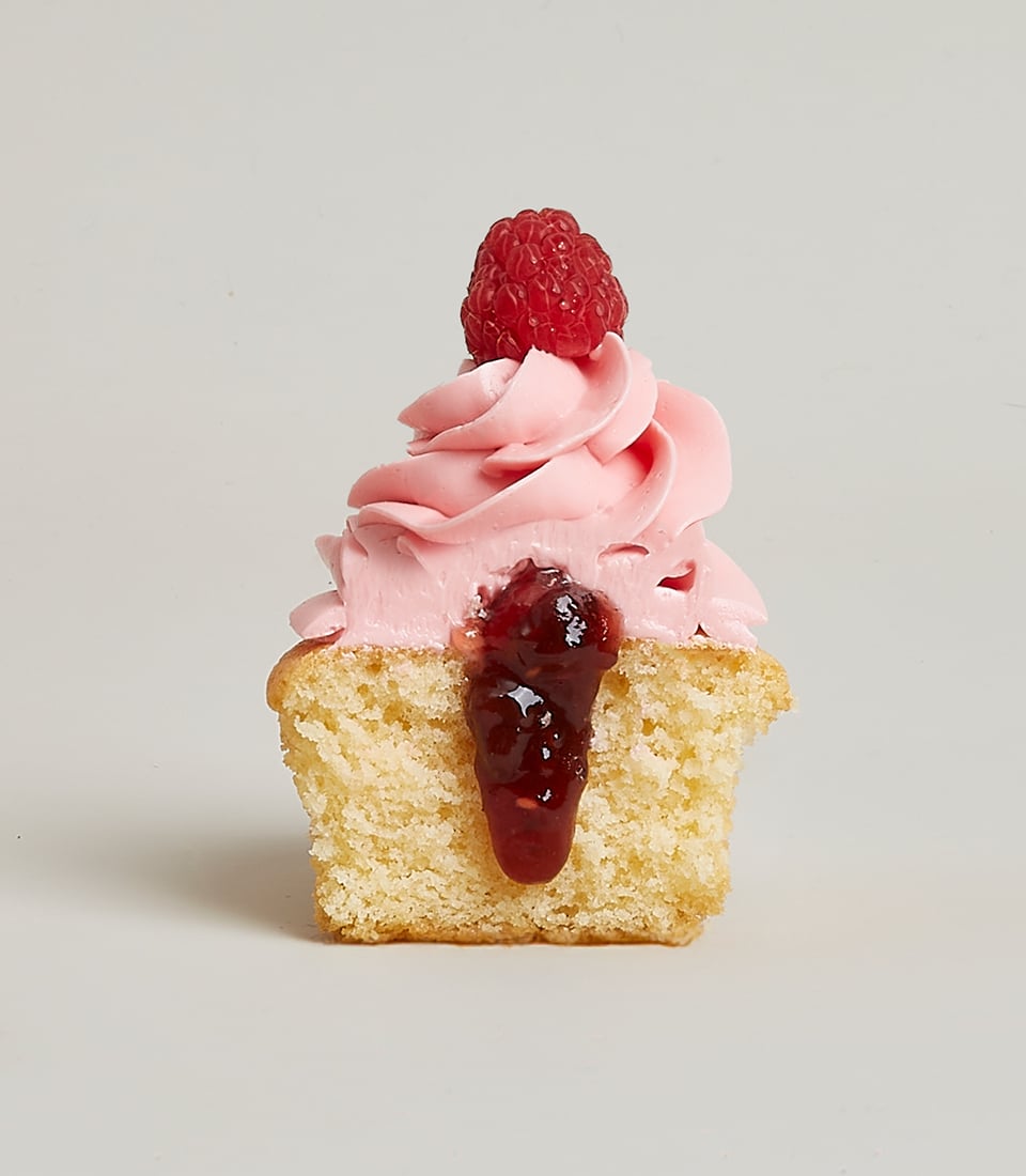 Light golden cupcake with raspberry jam center, pink frosting, and fresh raspberry on top