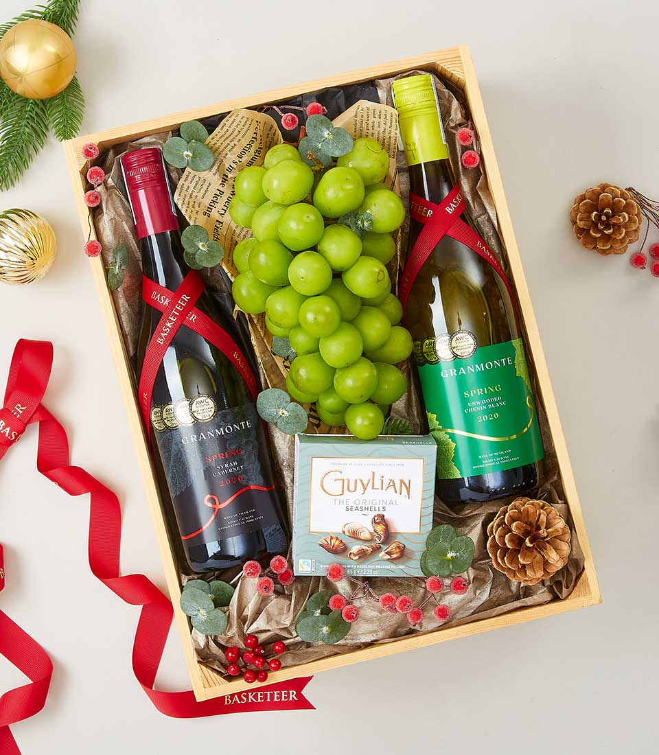 Premium wine and Guylian chocolate gift hamper with red and white wine, fresh grapes, and festive greenery in wooden crate