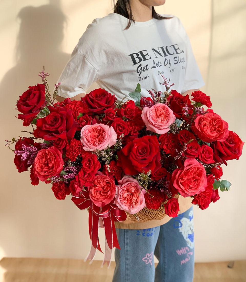 Scarlet and blush roses in woven basket with red carnations and purple filler flowers, tied with red striped ribbon