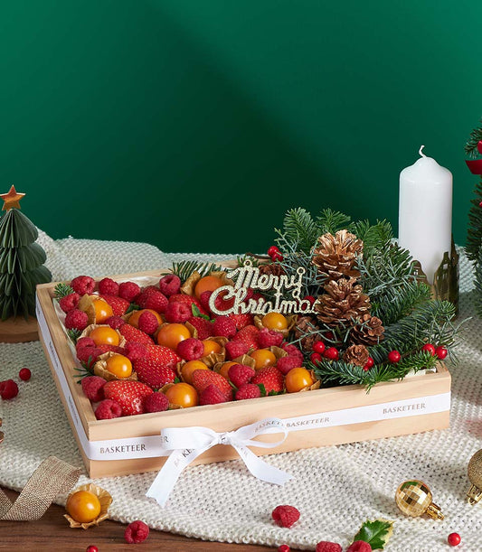 Festive wooden gift box filled with fresh strawberries, raspberries, and golden physalis with Christmas ornament and pine
