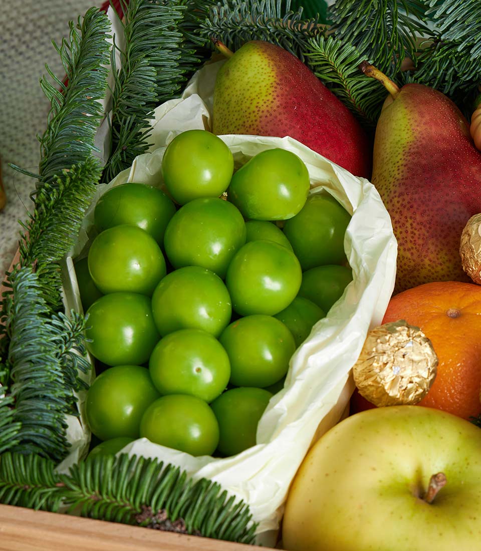 Festive wooden crate with fresh green plums, pears, apple, orange, Ferrero Rocher chocolate, and evergreen sprigs for