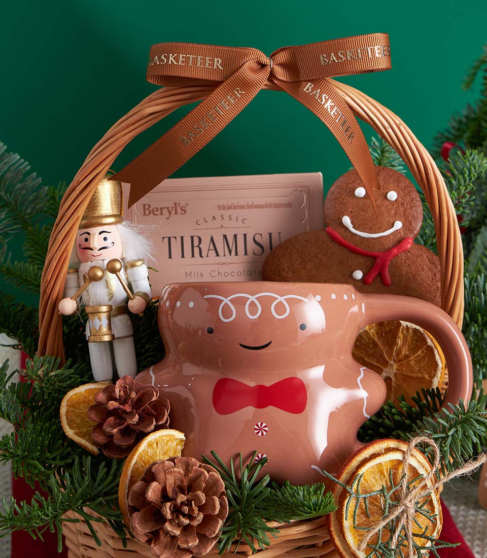Woven wicker Christmas gift basket with chocolate, shortbread, gingerbread cookies, ceramic mug, and nutcracker amid pine