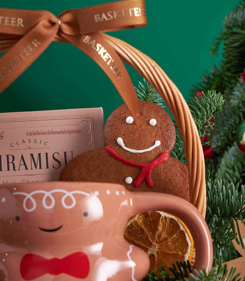 Festive Christmas gift basket with gingerbread cookies, tiramisu box, and ceramic gingerbread mug surrounded by pine branches