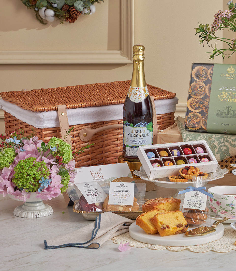 Luxurious wicker picnic basket filled with gourmet chocolates, artisan baked goods, sparkling beverage, and fresh floral